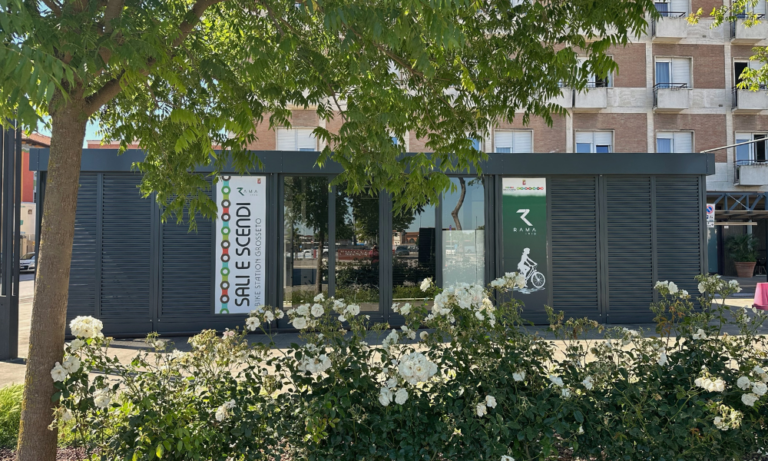 Ciclostazione Sali e Scendi di Grosseto, struttura moderna in metallo nero con pannelli a persiana, logo RAMA e scritta verticale, circondata da cespugli di ortensie bianche, con edifici residenziali in mattoni rossi sullo sfondo e un grande albero in primo piano.